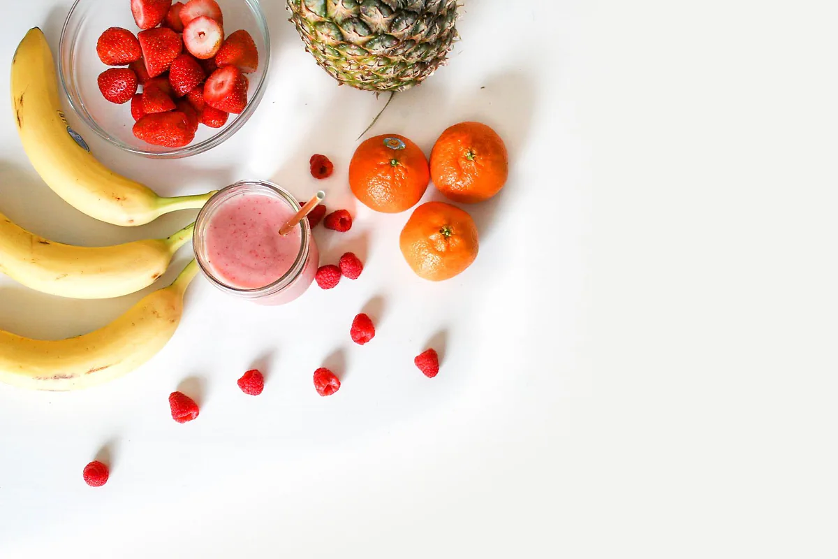 Bananas, strawberries, oranges, pineapple and raspberries around a pink smoothie